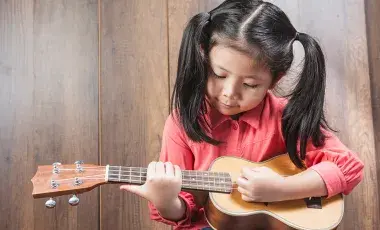 A young girl plays a ukulele in front of a wood-paneled wall.