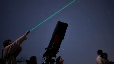 A person points to the night sky using a laser while standing next to a telescope. 